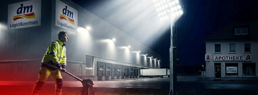 dm nutzt Apotheken-Blackout für Megaeröffnung Montage einer Person, die einen Strahler vor einem DM-Logistikzentrum einschaltet.