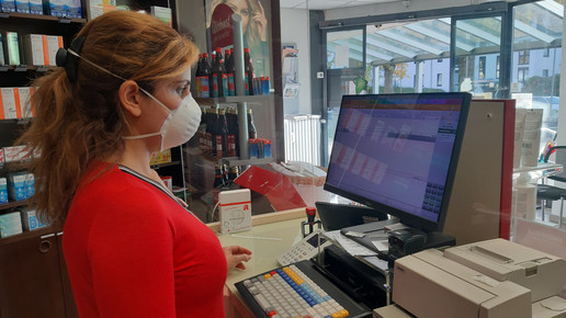 Apothekenmitarbeiterin mit Maske hinter dem HV, um sie rum eine hohe Plexiglaswand.