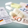 Retaxfalle Preisanker Foto: Tabletten, Geldscheine und Münzen neben einer Versichertenkarte auf einer Oberfläche.